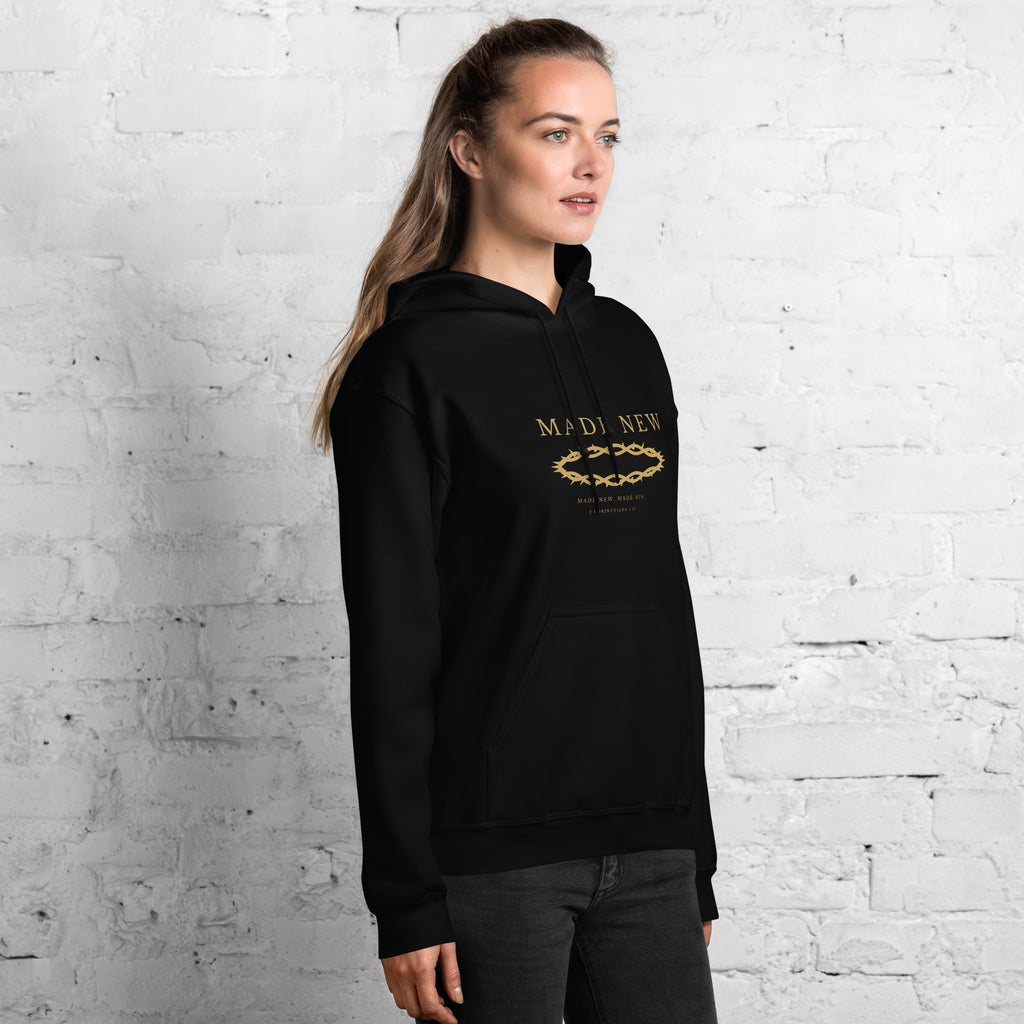 Woman wearing the Made New 2 Corinthians 5:17 black hoodie against a white brick wall, front view