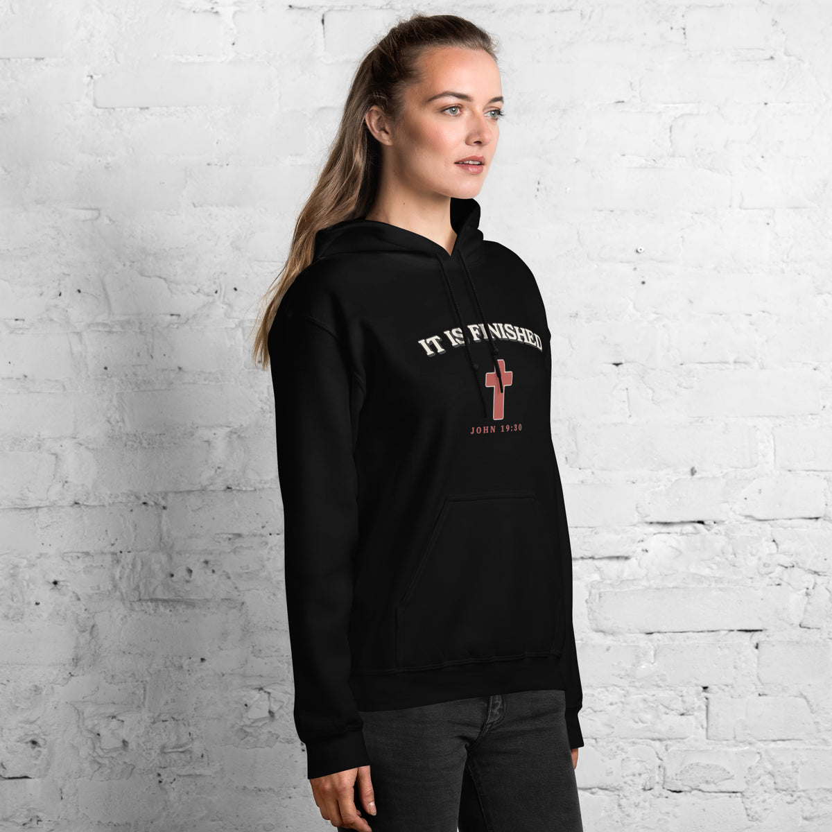Woman wearing the It Is Finished black hoodie against a white brick wall, side view